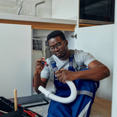 A plumber in blue overalls installs a new flexible drain pipe under a kitchen sink, with a toolbox beside him, providing expert plumbing repair and installation services.